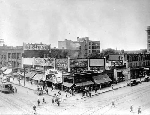 Historic black-and-white photo of a busy street corner with shops, signs, and pedestrians in an early 20th-century city.
