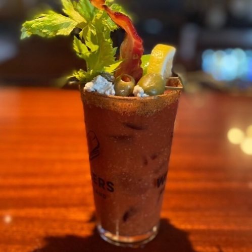A close-up of a garnished Bloody Mary cocktail in a glass, featuring celery, olives, and a slice of lemon.
