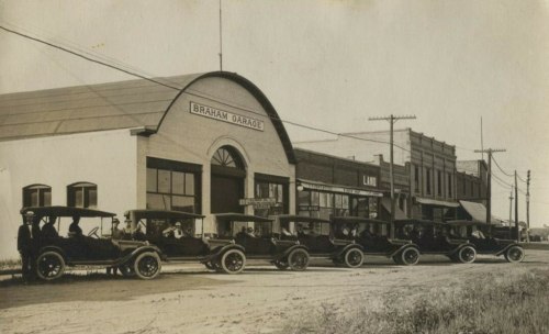 Historic photo of Braham Garage with several vintage cars parked in front, set on a dirt road.