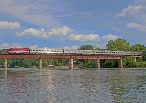 A train crosses a bridge over a river, surrounded by green trees and a blue sky with fluffy clouds.