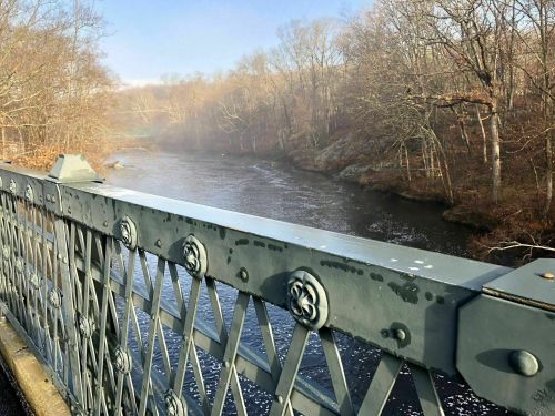 A misty river flows beneath a metal bridge, surrounded by bare trees in a tranquil landscape.