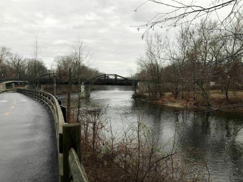 A calm river flows beside a path, with a bridge in the background and bare trees lining the shore under a cloudy sky.