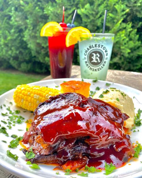A plate of barbecue ribs with corn, mashed potatoes, and garnished with herbs, alongside colorful cocktails.