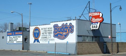 A pizzeria building with a large sign reading "Buddy's Pizza" and a mural advertising local news and entertainment.