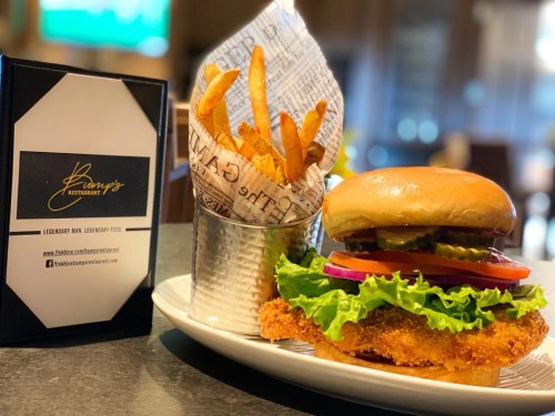 A crispy chicken burger with lettuce, tomato, and pickles, served with a side of golden fries in a metal cup.