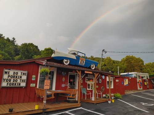 A colorful diner with a vintage car on the roof, under a rainbow and surrounded by green hills.