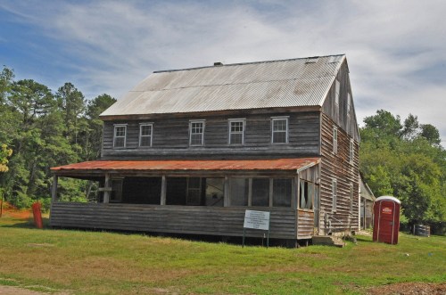 A weathered two-story wooden building with a metal roof, surrounded by grass and trees, featuring a portable restroom nearby.