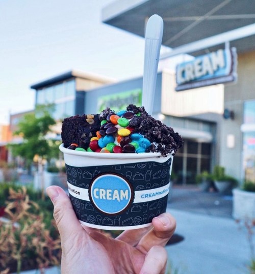 A hand holds a cup of ice cream topped with cake, colorful candies, and a spoon, with a shop in the background.