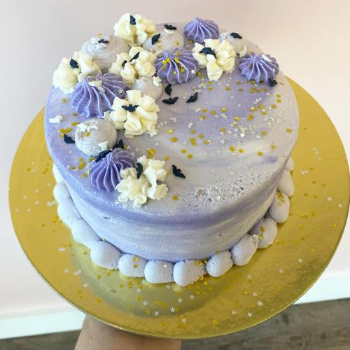A round lavender cake decorated with purple frosting, white flowers, and gold sprinkles on a gold cake board.