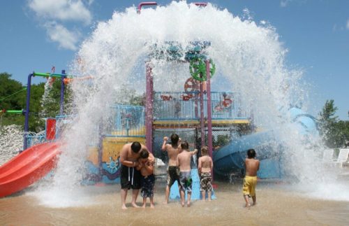 Children play in a splash pad, water cascading down from above, with colorful slides in the background on a sunny day.