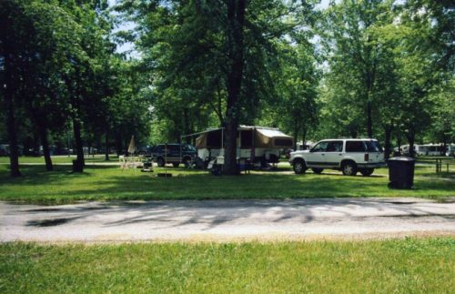 A campsite with two vehicles parked near a pop-up camper, surrounded by trees and grassy areas.