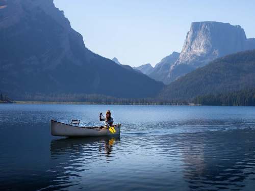 A person paddles a canoe on a calm lake surrounded by mountains under a clear blue sky.