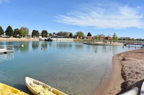 A serene lake scene with kayaks on the shore, trees, and a park area in the background under a clear blue sky.