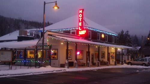 A cozy hotel and café adorned with colorful lights, surrounded by snow and mountains on a cloudy winter day.
