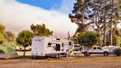 A camper trailer parked under tall trees at a campsite, with a truck nearby and tents in the background.