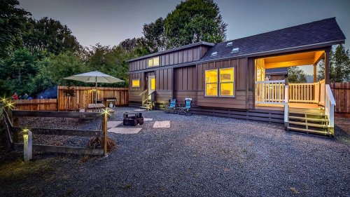 A cozy modern home with warm lights, surrounded by a gravel yard and a patio area with chairs and an umbrella.
