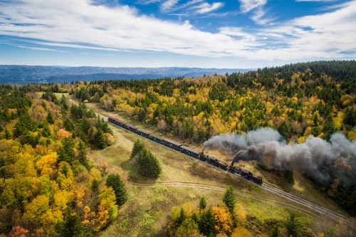 A steam train travels through a colorful autumn landscape, surrounded by trees and rolling hills under a blue sky.