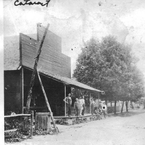 Black and white photo of a group of people standing on a porch of a wooden building, with trees in the background.