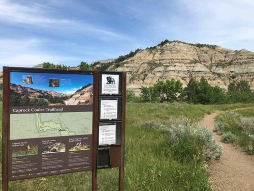 Trailhead sign for Caprock Coulee, with a scenic view of layered hills and a grassy path leading into the landscape.