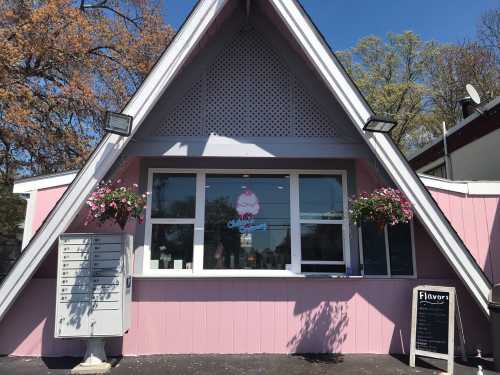 A pink A-frame ice cream shop with flower baskets, a menu board, and a window displaying ice cream offerings.