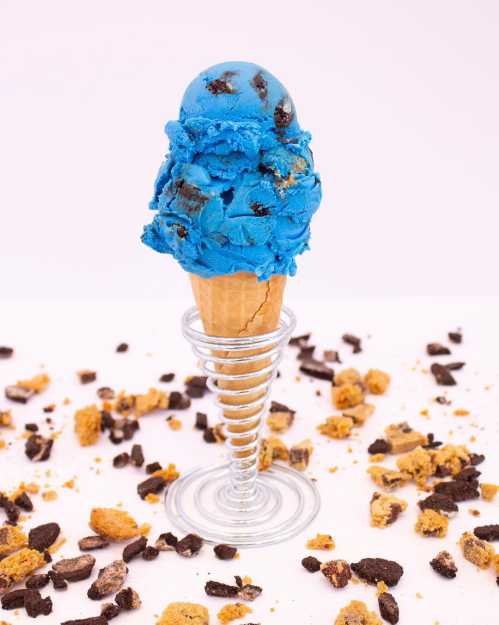 A blue ice cream cone topped with cookie pieces, surrounded by scattered cookie crumbs on a white background.