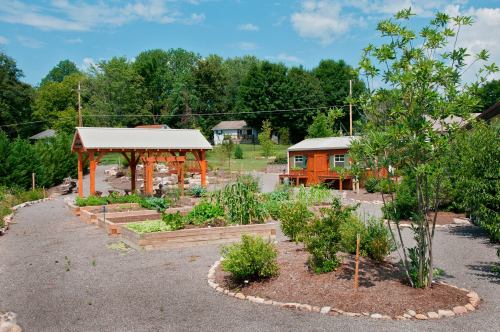 A vibrant community garden featuring raised beds, a gazebo, and two small wooden buildings surrounded by greenery.
