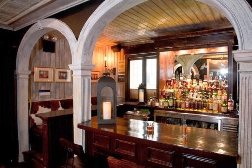 A cozy bar with wooden arches, a polished counter, and a well-stocked shelf of various liquor bottles in the background.