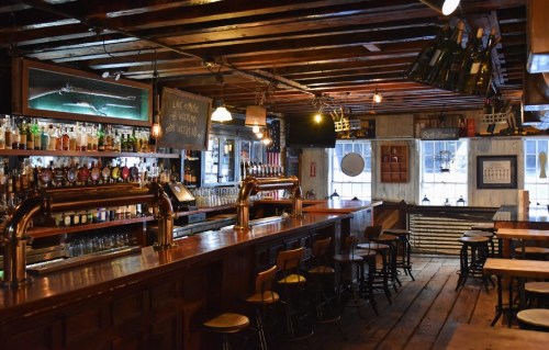 A cozy bar interior with wooden beams, a long counter, shelves of liquor, and warm lighting.