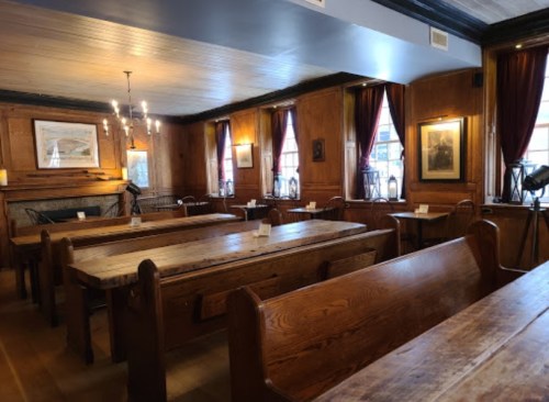 A cozy, wooden dining room with long tables, chairs, and warm lighting from chandeliers and windows.
