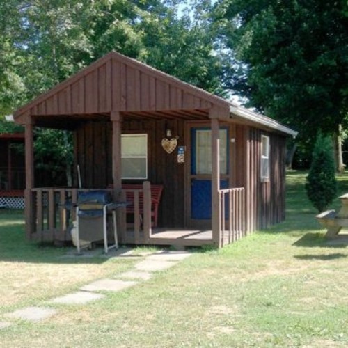 A small wooden cabin with a porch, surrounded by trees and grass, featuring a heart decoration on the front.