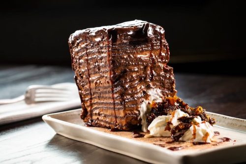 A tall slice of chocolate cake drizzled with chocolate sauce, served with whipped cream on a plate.