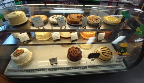 A display case filled with various cakes and pies, each labeled with names and prices.