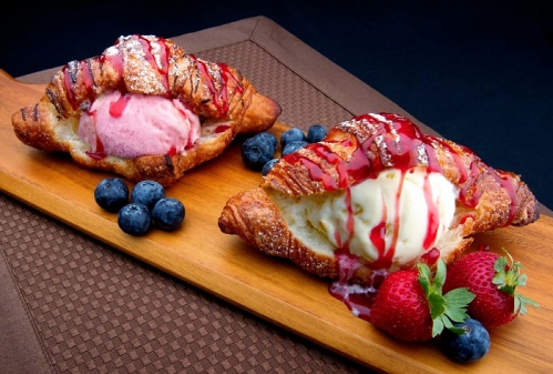 Two croissants filled with ice cream, drizzled with sauce, surrounded by strawberries and blueberries on a wooden platter.