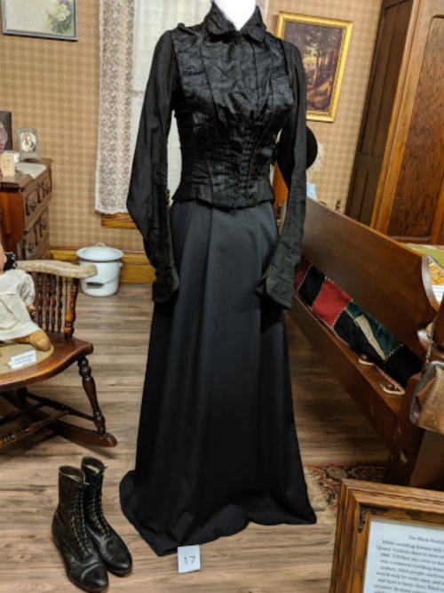 A vintage black dress with a fitted bodice and long skirt displayed in a museum setting, alongside black boots.