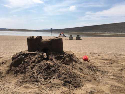 A sandcastle on a beach with a lake in the background, and people relaxing under an umbrella nearby.