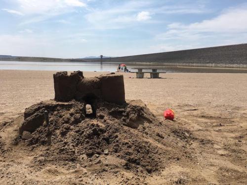 A sandcastle on a beach with a lake in the background, and people relaxing under an umbrella nearby.