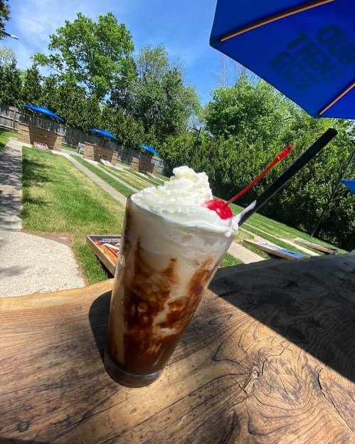A tall glass of chocolate milkshake topped with whipped cream and a cherry, set outdoors with green trees in the background.
