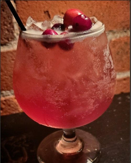 A refreshing cocktail in a glass, filled with ice and garnished with cranberries, against a brick wall background.