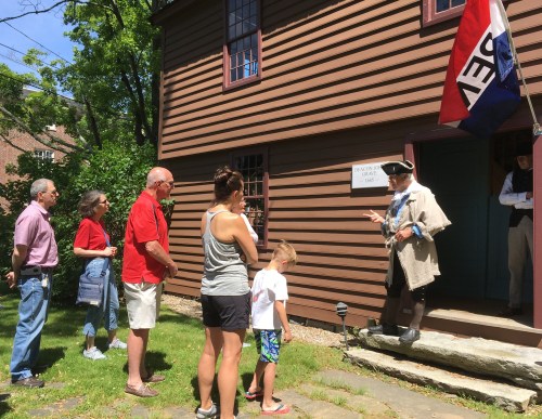 A historical guide speaks to a small group outside a brown wooden building, with a flag flying nearby on a sunny day.