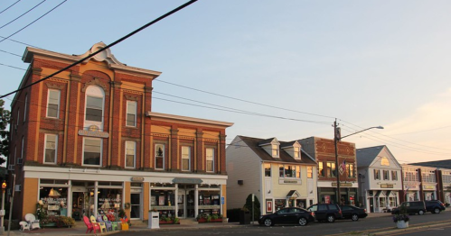 Historic brick buildings line a street, featuring shops and colorful chairs outside, under a clear sky at sunset.