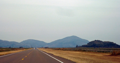 A wide, empty road stretches towards distant, hazy mountains under a gray sky. Fields line the sides of the road.