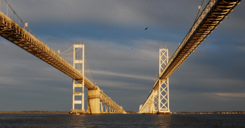 A view of two bridges spanning a body of water, with dramatic clouds and sunlight illuminating the structures.