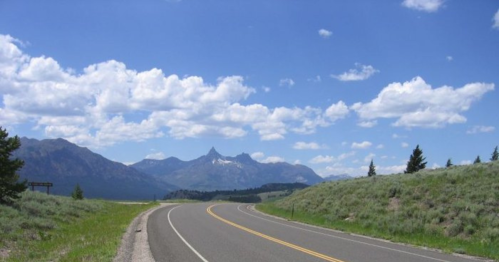 A winding road leads through green hills with mountains and a blue sky filled with clouds in the background.