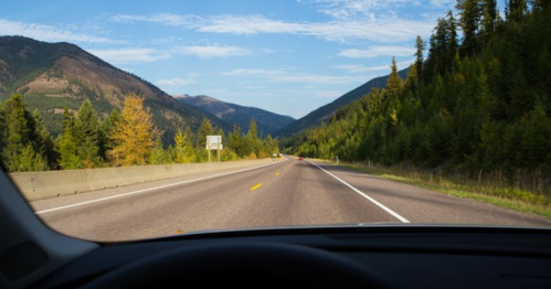 View from a car dashboard on a winding road surrounded by mountains and trees under a clear blue sky.