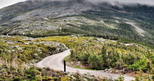 Winding road through a mountainous landscape with greenery and rocky terrain under a cloudy sky.