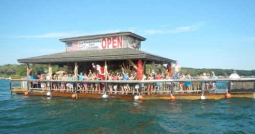 A lively waterfront bar with a crowd of people celebrating, surrounded by water and trees under a clear blue sky.