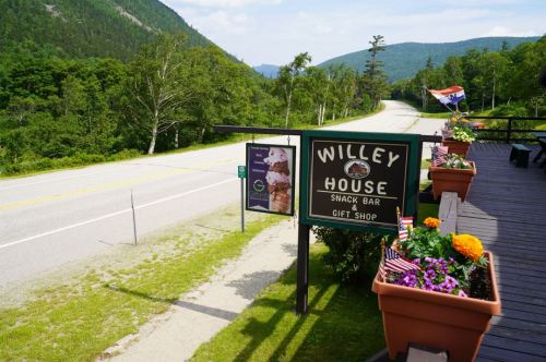 Sign for Willey House Snack Bar and Gift Shop, with colorful flower pots and a scenic view of a road and mountains.