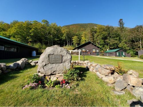 A large rock with a plaque in a grassy area, surrounded by flowers, with cabins and mountains in the background.