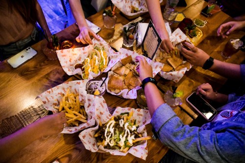 A table filled with various dishes, including tacos, fries, and drinks, with hands reaching for the food.