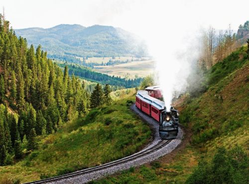 A vintage steam train winds through lush green hills and mountains, with smoke billowing against a scenic backdrop.